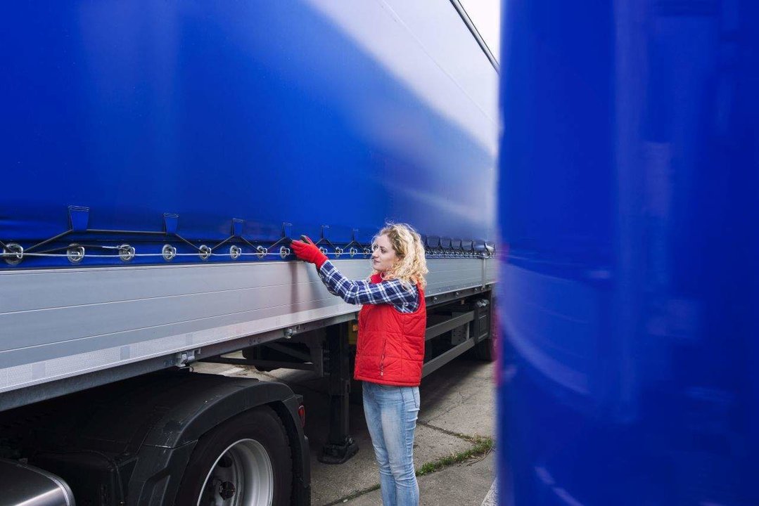 Female truck driver checking vehicle and tightening truck tarpaulin.